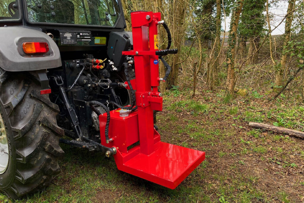 Red hydraulic farm implement attached to a tractor, with hoses and control valves visible in a woodland setting on uneven ground.