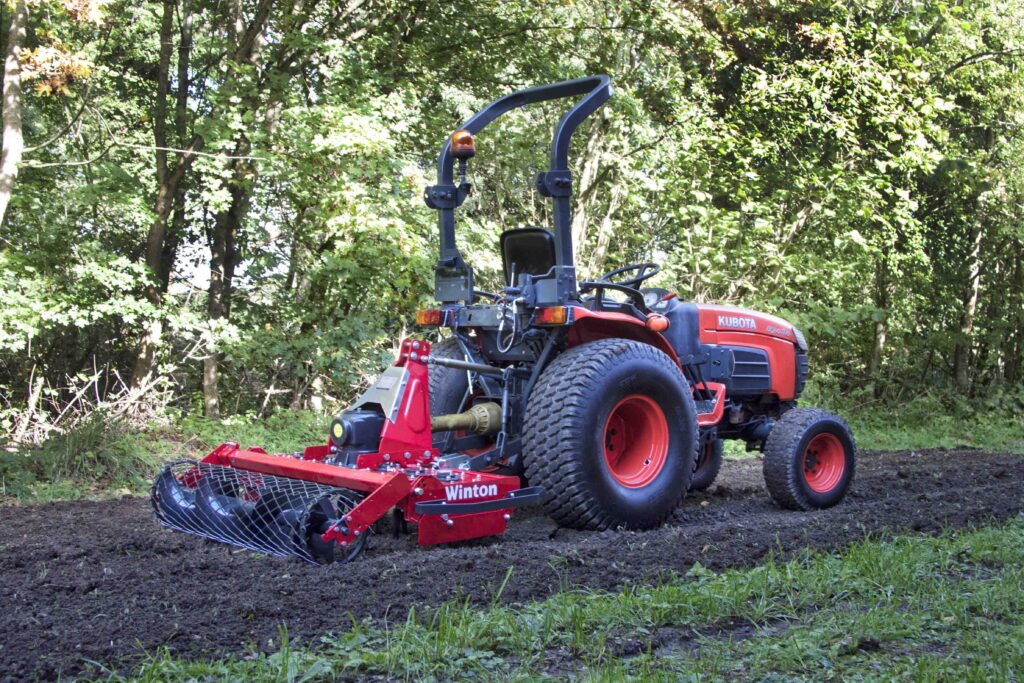 Red Kubota tractor with a large attached implement resting on a dirt patch in a wooded yard area.