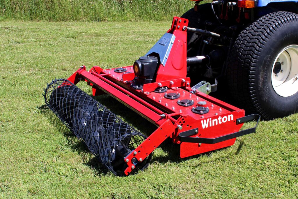 Red agricultural implement attached to a tractor on a grassy field, with a large tire visible in the background. The machinery shows bolts and a blue support plate.