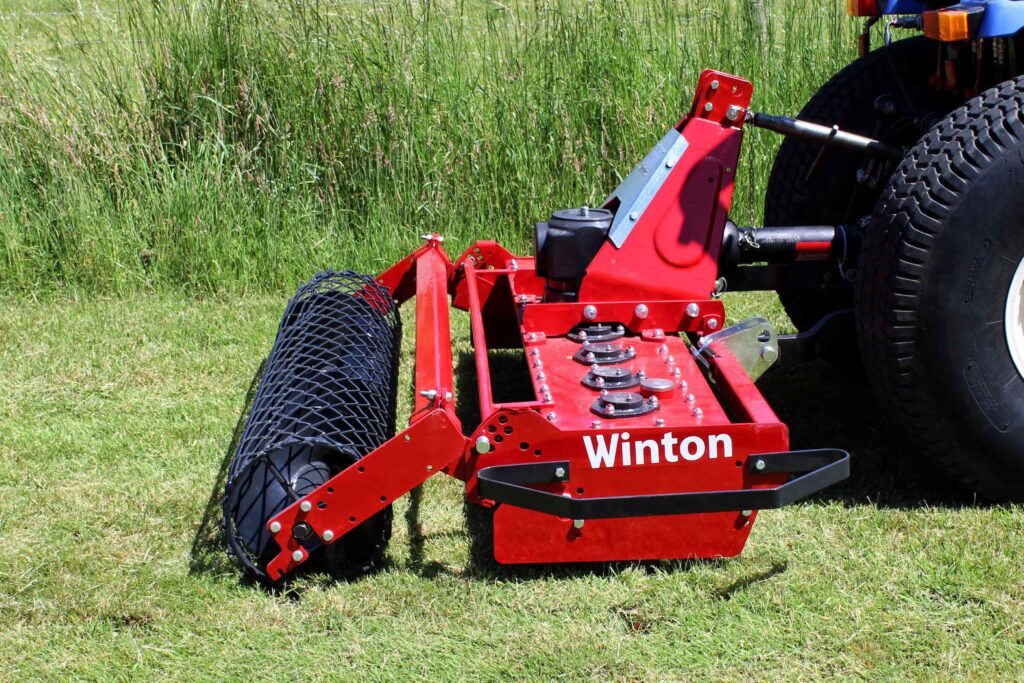 Red farm equipment attached to a tractor sits on a grassy field, with a large tire visible on the right and the name 'Winton' on the frame.
