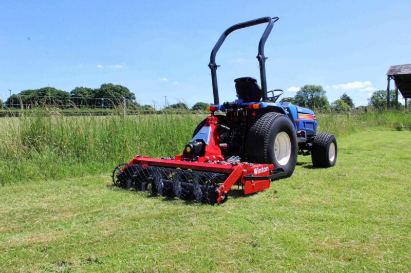 Blue tractor with a red tillage implement attached in a sunny rural field.