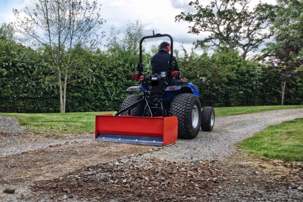 Person wearing orange hearing protection sits on a blue tractor with a large red front blade on a gravel path, surrounded by trees and bushes in a park-like setting.