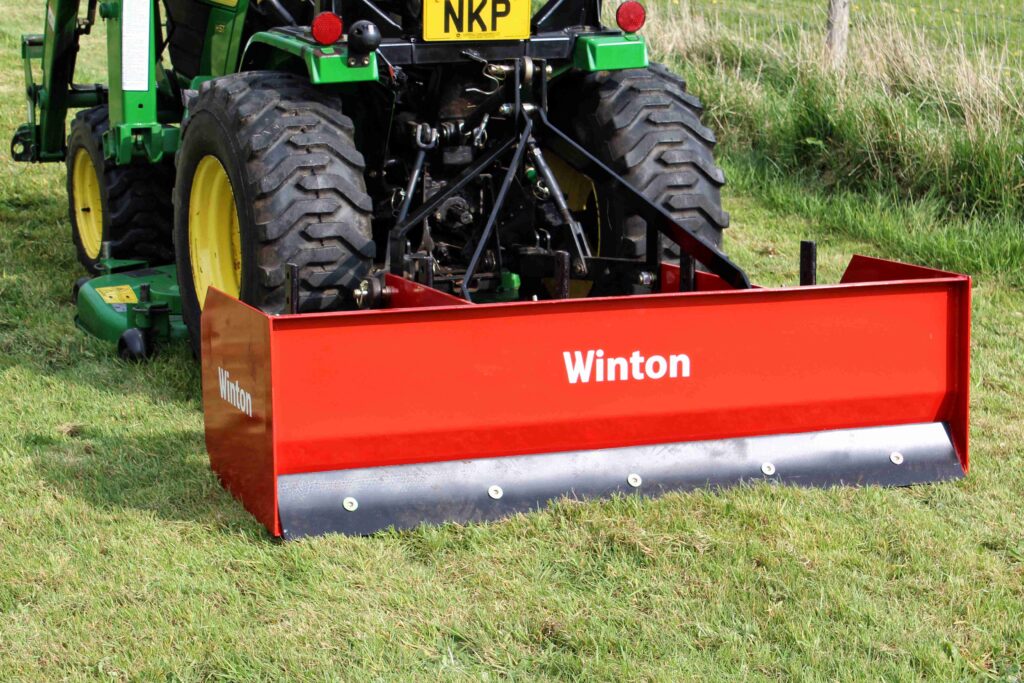 Rear view of a green tractor with large tires pulling a bright red front-mounted implement labeled 'Winton' in a grassy field.