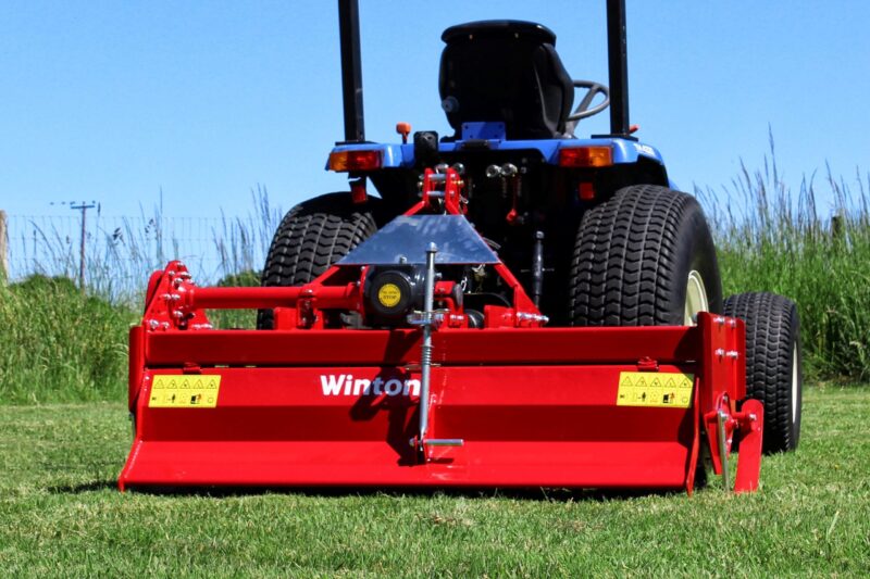 Red farming implement attached to a blue tractor on a grassy field, with large rear tires visible and warning labels on the equipment.