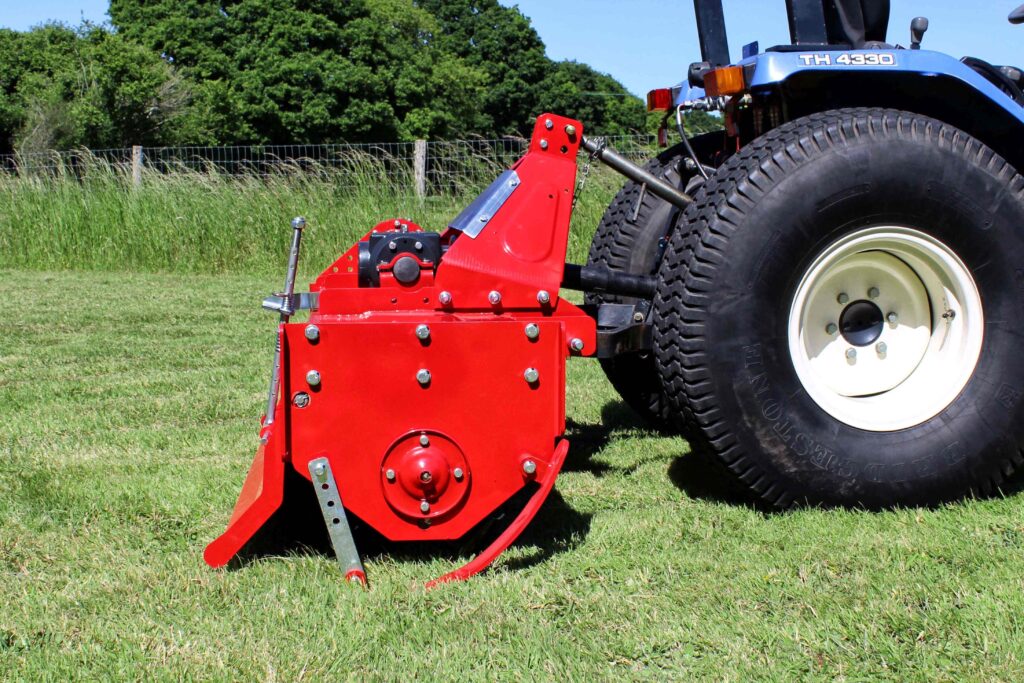 Red farming implement hitched to a blue tractor in a grassy field with a fence and trees in the background.