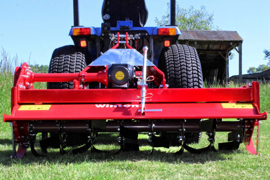Rear view of a red farm implement with large dual tires on a trailer, showing hitch and warning labels, on a grassy field with a wooden shed in the background.