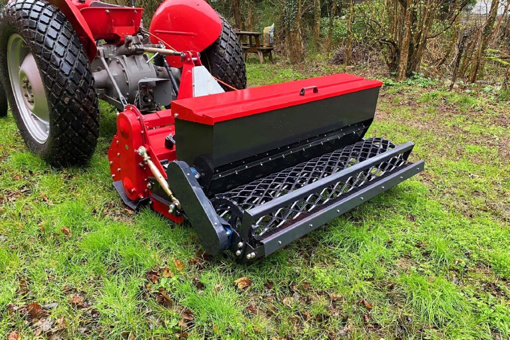 Red agricultural implement attached to a tractor in a grassy, wooded area, featuring a black metal rake attachment in front.