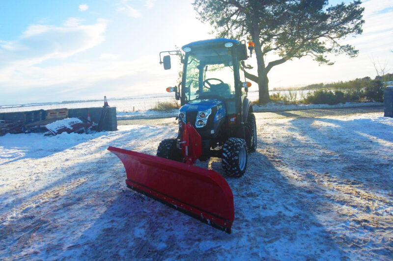 Blue tractor with a red snow plow attached sits on a snow-covered lot beside a tree and a body of water in the background