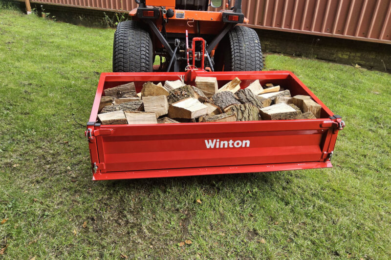 Red trailer filled with chopped firewood on a grassy lawn behind a vehicle. Brand ‘Winton’ visible on the trailer side.