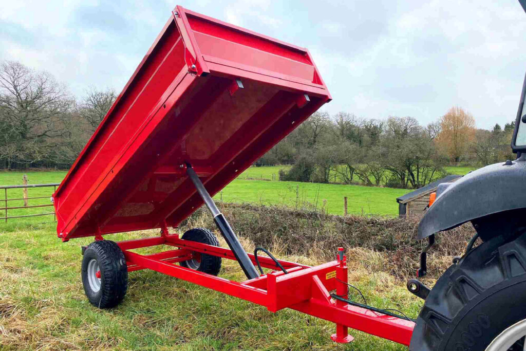 Red tipping trailer raised on a hydraulic cylinder beside a tractor in a grassy field.