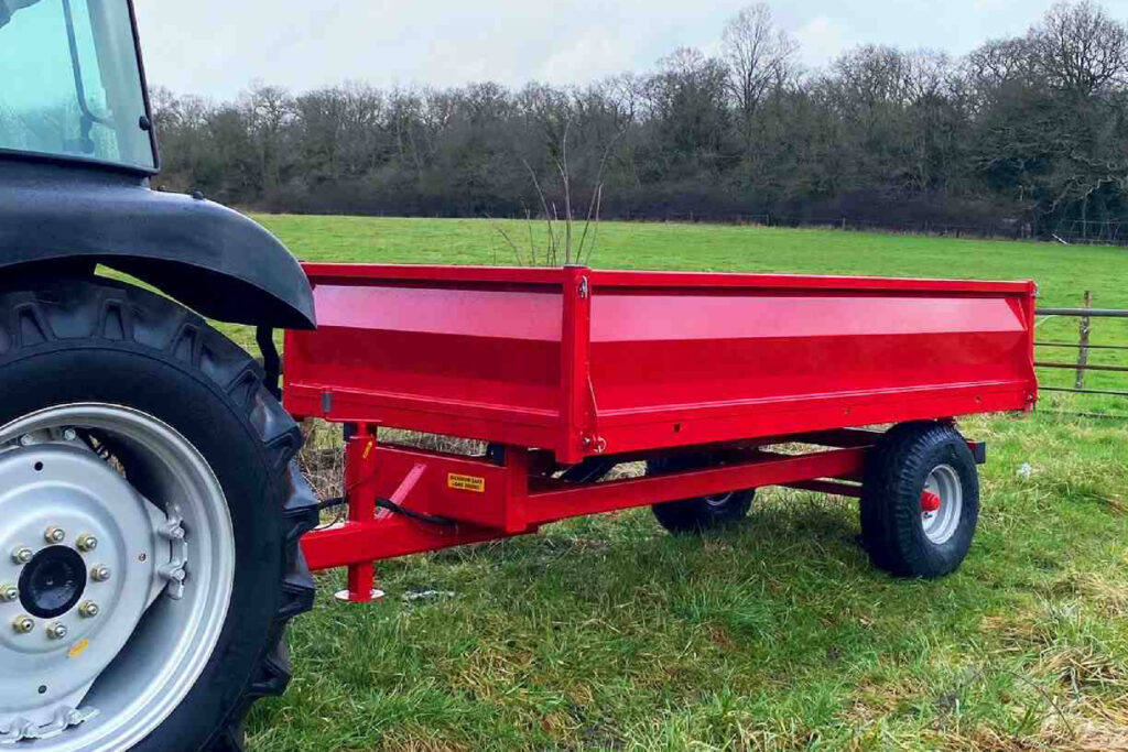 Front left wheel and fender of a dark tractor attached to a bright red trailer in a grassy field with trees in the background