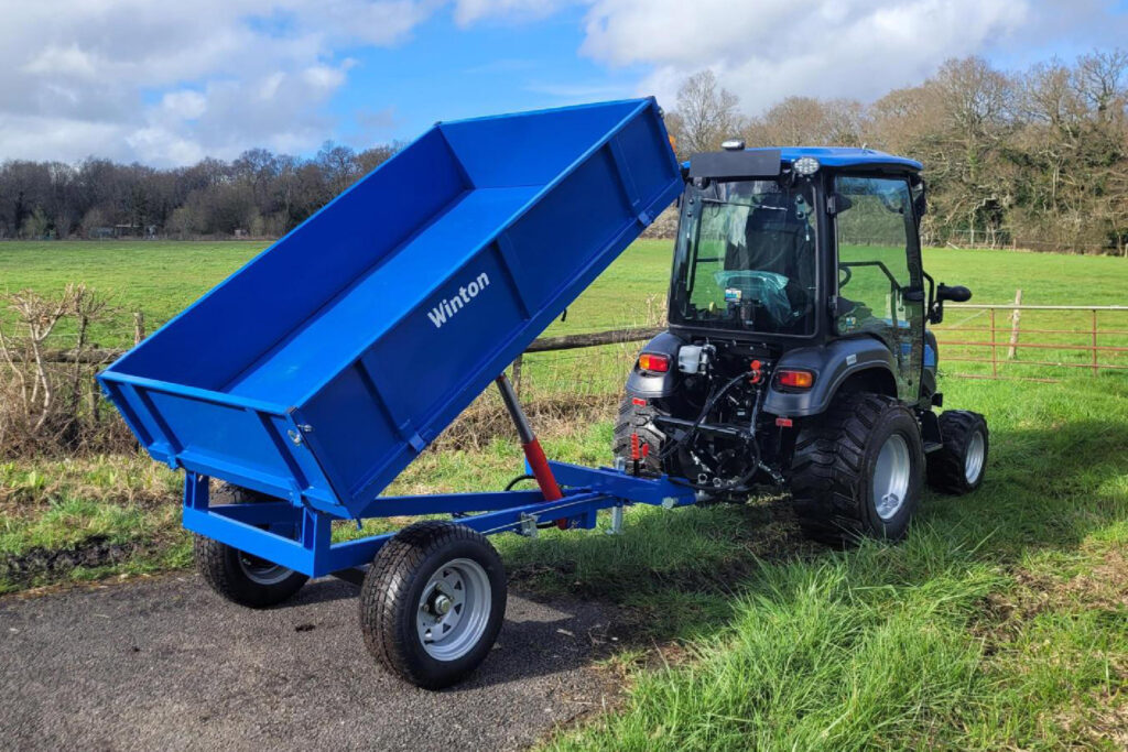 Blue tipping trailer raised on a compact tractor in a grassy field, ready to dump its load.