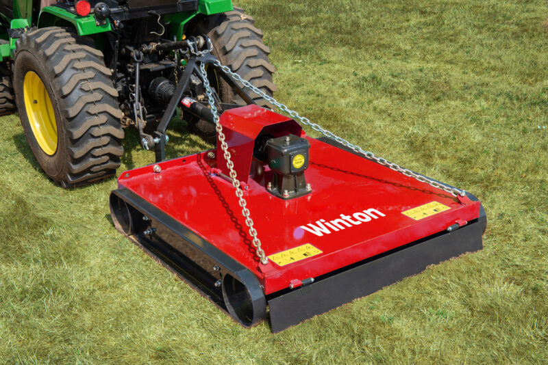 Red mower attachment hitched to a green tractor on a grassy field, ready for mowing; chains secure the hitch and 'Winthon' branding visible on the deck.