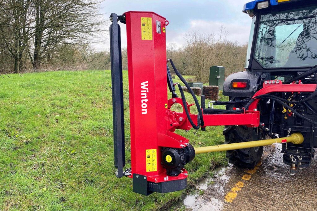 Red agricultural implement attached to a tractor in a grassy field, with hoses and warning labels visible.