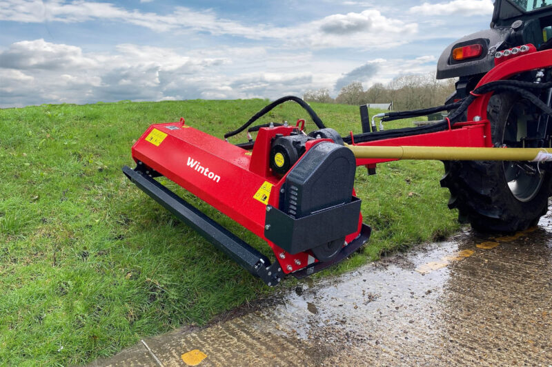 Red agricultural mower attachment linked to a tractor on a grassy field under a cloudy sky.